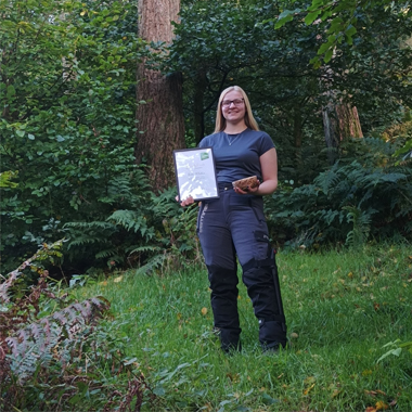 Photo of student Ellie Watton standing in forest holding Arboricultural Association Student of the Year award